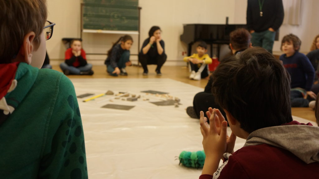 deux enfants, de dos, observent des objets sonores placés au centre d'un groupe d'élèves